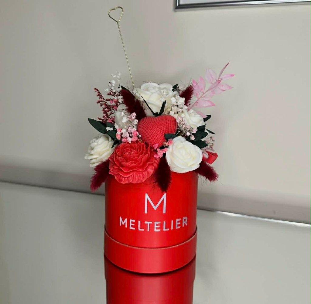 A red decorative container filled with an arrangement of artificial flowers including white roses, pink foliage, and a red heart, labeled 'MELTELIER'.